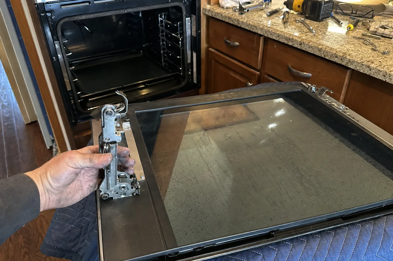 A built-in oven door lying glass-up on a quilted blanket while a hand holds the hinge assembly clear of it