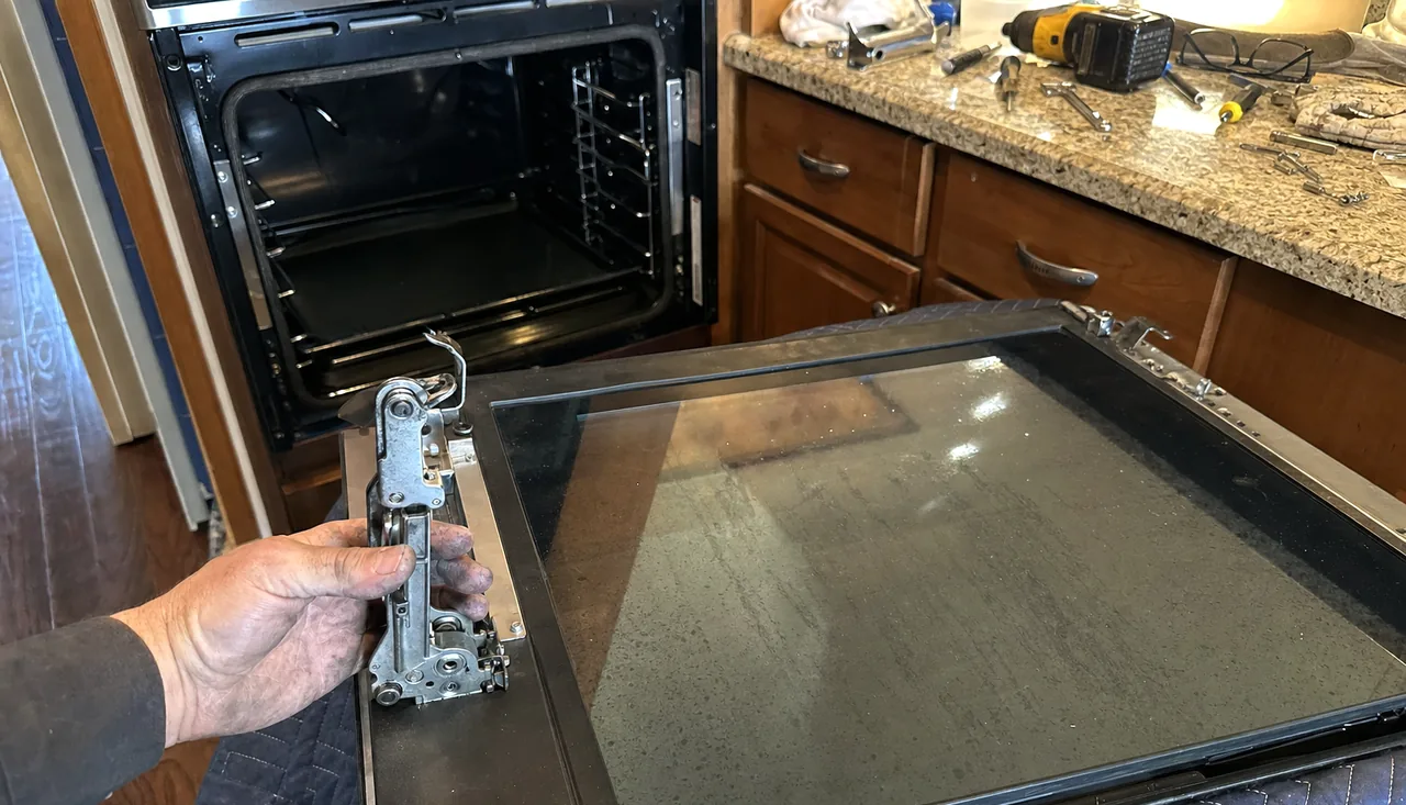 A hinge assembly held in a working hand above an oven door, spanners and drivers spread on the counter behind