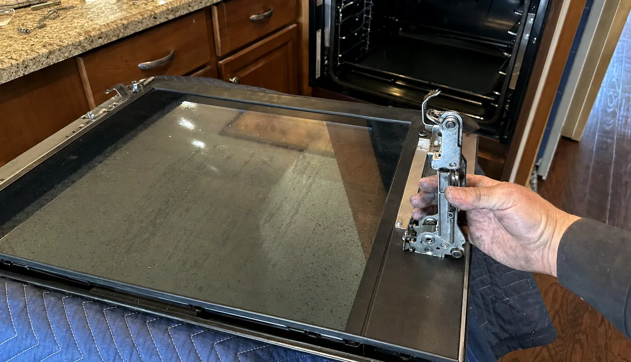 An oven door on a moving blanket with the hinge arms extended and a screwdriver resting alongside