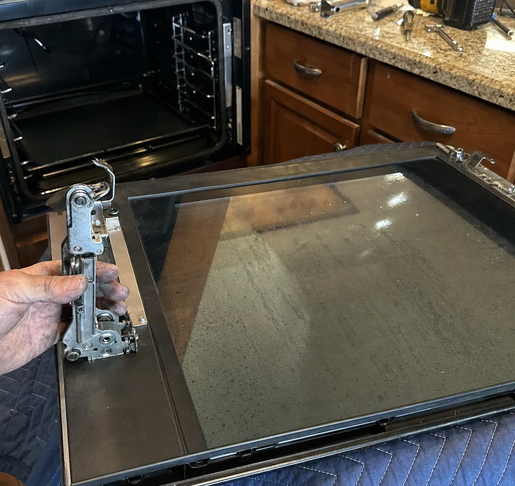An open built-in oven with bare rack guides beside a granite counter, while a hand steadies the hinge of its removed door laid flat on a blue moving blanket